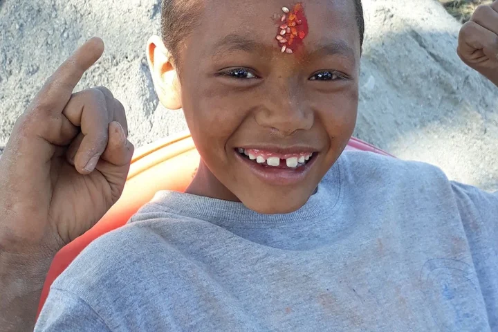 A boy grinning at the camera, a tika of rice and vermilion on his forehead