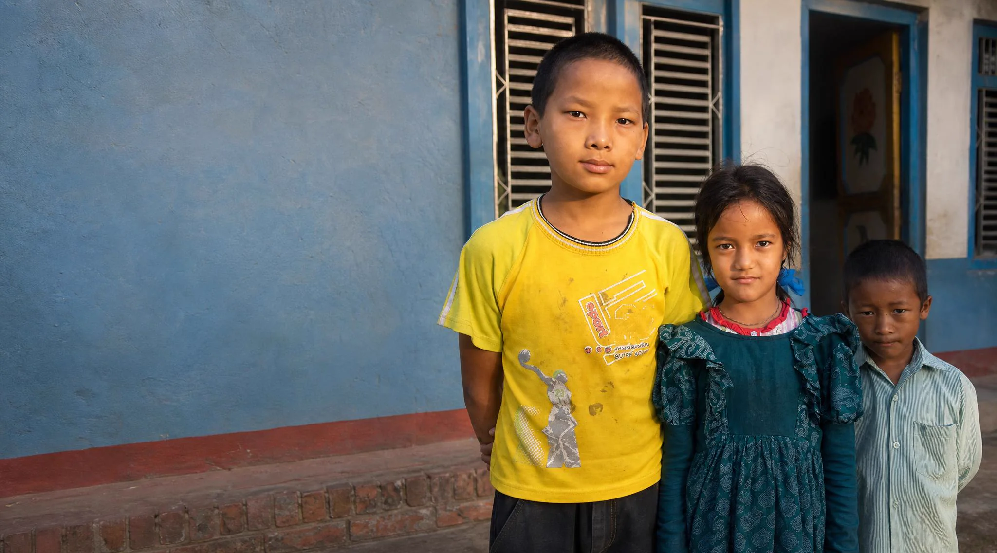 Three children stand against the blue wall of their school in rural Nepal, looking at the camera