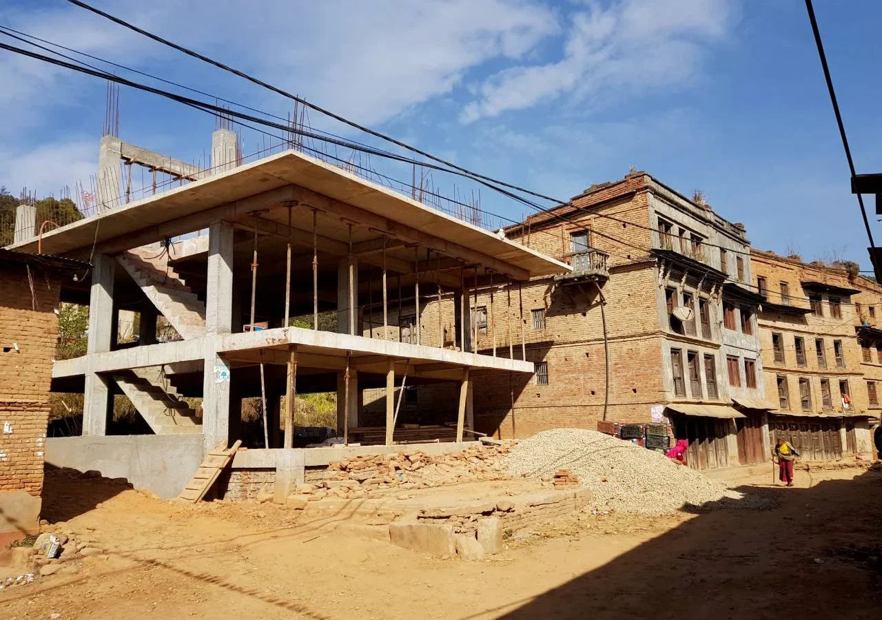 A reinforced concrete frame rising between older brick houses in a Nepali village