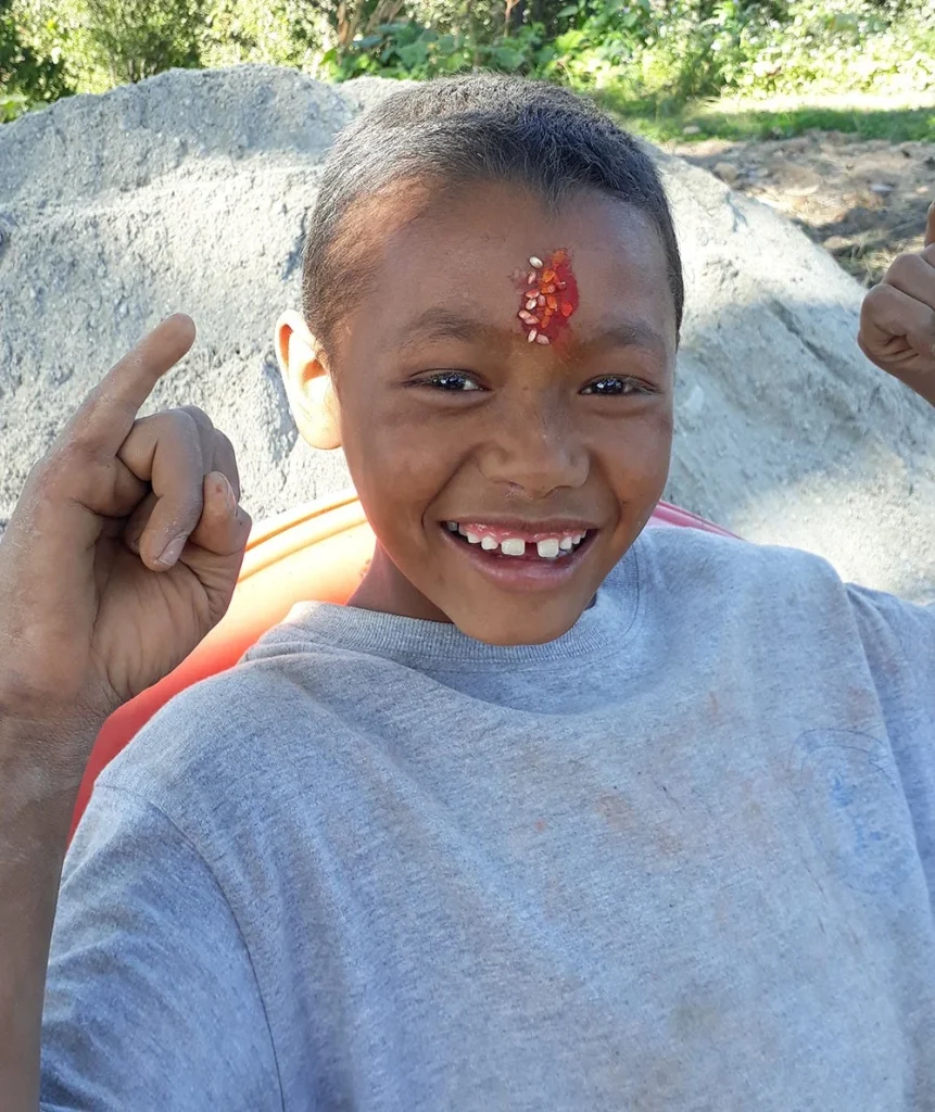 A boy grinning at the camera, a tika of rice and vermilion on his forehead