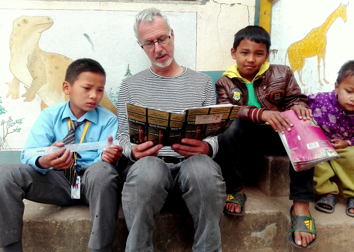 Alain Rouveure reads with children in front of a painted mural wall at a school in Nepal, one boy holding a ruler and another holding a new exercise book