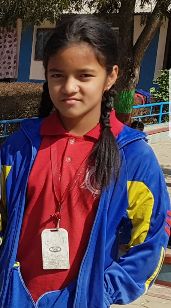 A girl in a red and blue Namo Buddha school uniform, wearing her school ID card, standing in the school yard