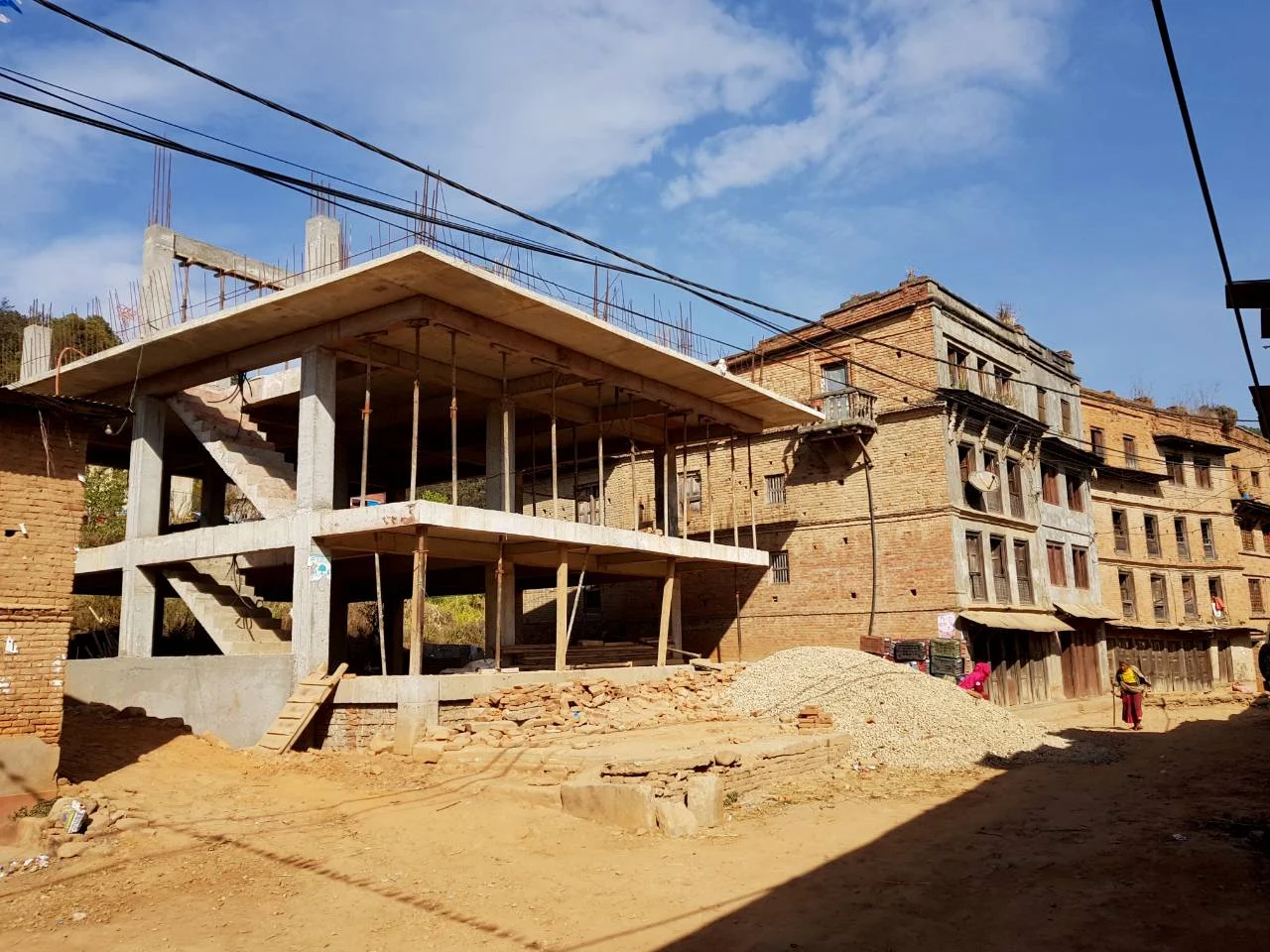 A reinforced concrete pillar-and-slab frame under construction on a village street in Nepal, standing beside older brick houses, with a pile of gravel and a woman walking past